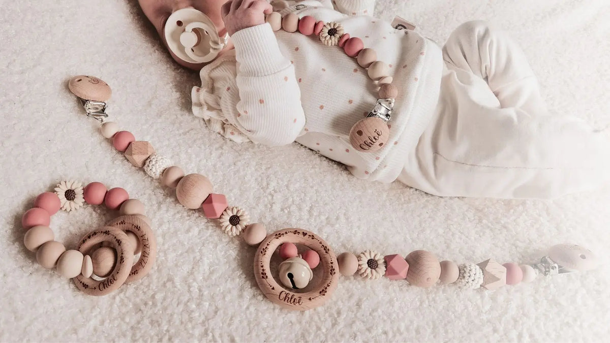 Baby met een gepersonaliseerde roze bijtring, wagenspanner en speenkoord, gegraveerd met naam.