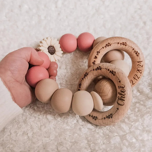 Gepersonaliseerde roze bijtring in babyhandje, gegraveerd met hartjes, takjes, naam en geboortedatum.